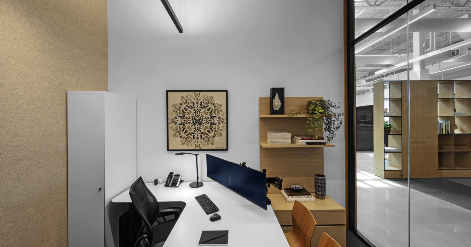 Minimalist office space boasting a sleek white desk with a desktop setup, ergonomic black chair, and decorative plants juxtaposed against a monochromatic color scheme, accentuated by geometric wall art and framed by linear suspended lighting. The glass partition reflects an open-plan environment beyond.