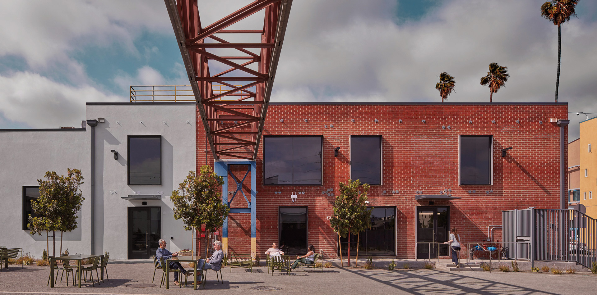 An urban oasis where contemporary architecture meets industrial design, featuring locals enjoying an outdoor seating area under the geometric embrace of an overhead red steel structure, with a serene backdrop of blue skies and palm trees.