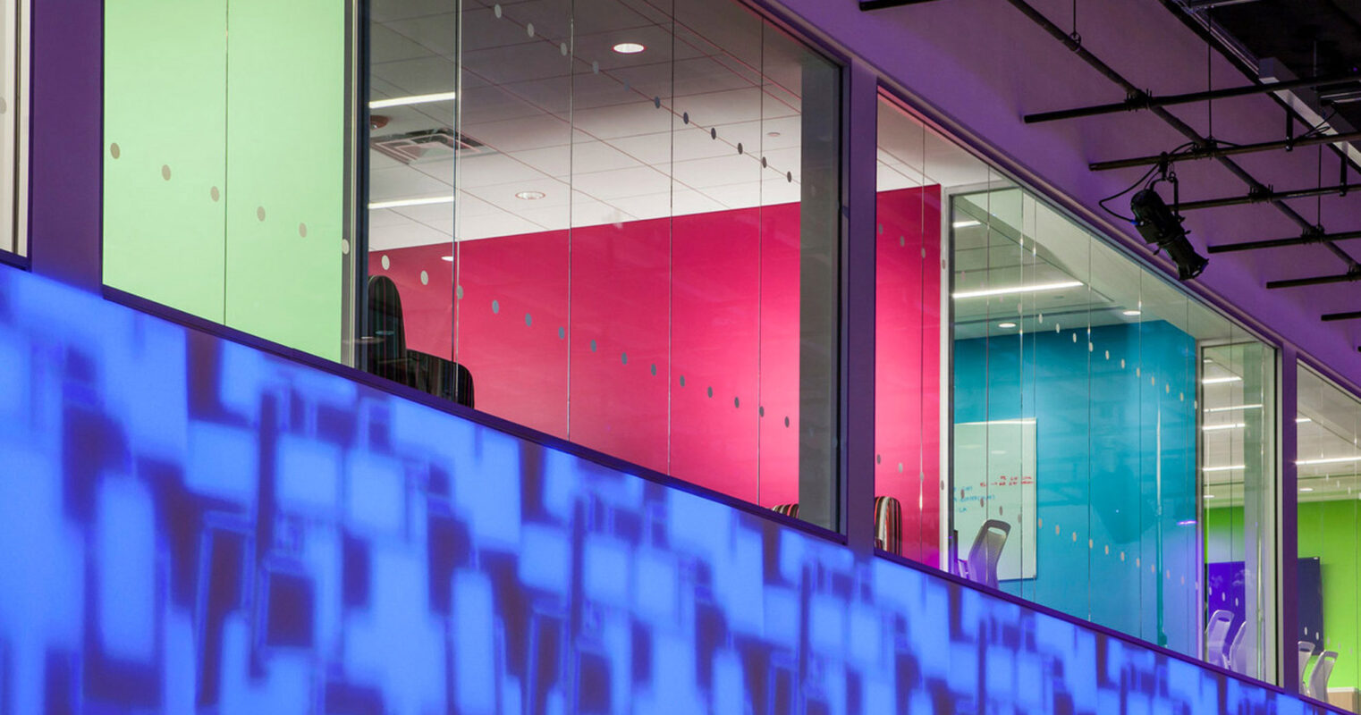 Vibrant interior featuring a glass-walled mezzanine with translucent panels in gradient shades of pink and green. The space is accentuated by blue LED lighting casting geometric shadows, creating a dynamic and modern atmosphere.