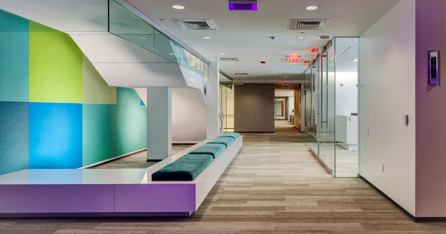 Modern office hallway featuring muted gray carpeting, with a vibrant geometric accent wall in green and blue hues, complemented by sleek white built-in seating and frosted glass partitions, creating a contemporary and welcoming workspace.