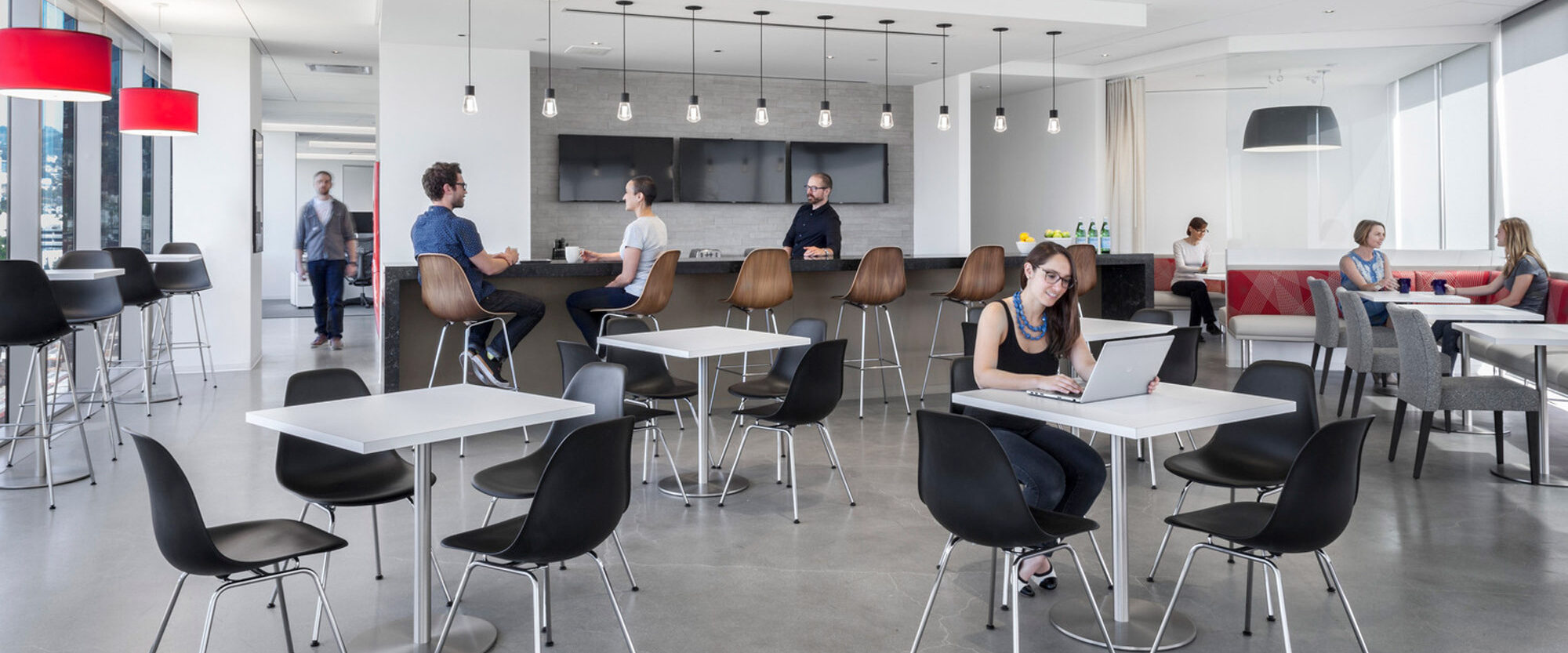 Modern office break room featuring a minimalist design with monochromatic tones, accented by red pendant lights. A mix of high-top tables with stools and standard height seating provides variety, while natural light filters in, enhancing the sleek, functional space. People engage casually and work on laptops.