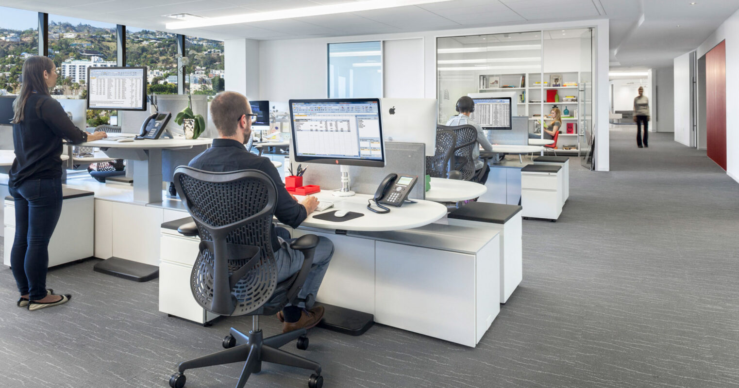 Modern open-plan office with sleek, white desks and ergonomic black chairs. Employees work at computers with dual monitor setups. The room features a clean design with natural light and an uncluttered shelving unit. A city landscape is visible through floor-to-ceiling windows.