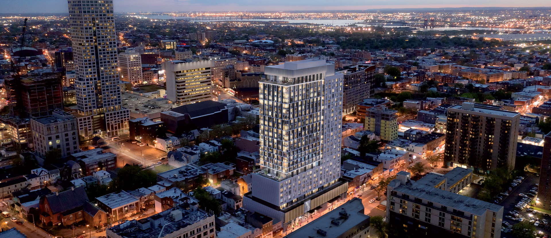 Twilight cityscape with high-rise buildings illuminated from within, showcasing varied architectural styles and grid street layout, highlighting urban density and mixed-use development.