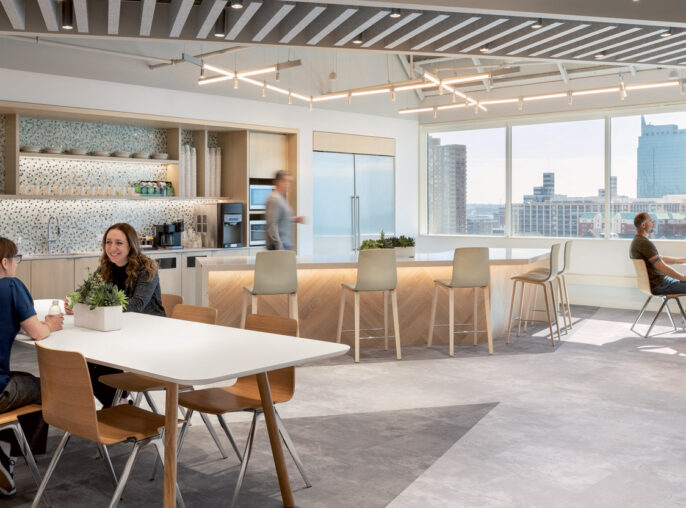 Modern office break room with natural wood tables, light gray flooring, and suspended linear LED lighting. A kitchenette with mosaic backsplash and high stools overlooks a cityscape through large windows. Employees casually engage, fostering a collaborative atmosphere.
