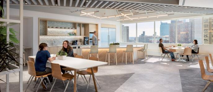 Modern office break room with natural wood tables, light gray flooring, and suspended linear LED lighting. A kitchenette with mosaic backsplash and high stools overlooks a cityscape through large windows. Employees casually engage, fostering a collaborative atmosphere.