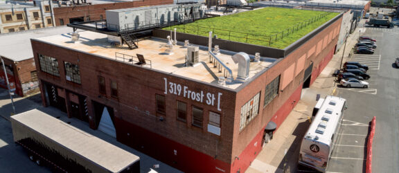 Rooftop urban garden above a brick industrial building, showcasing modern sustainable design with lush greenery against an expansive cityscape background.