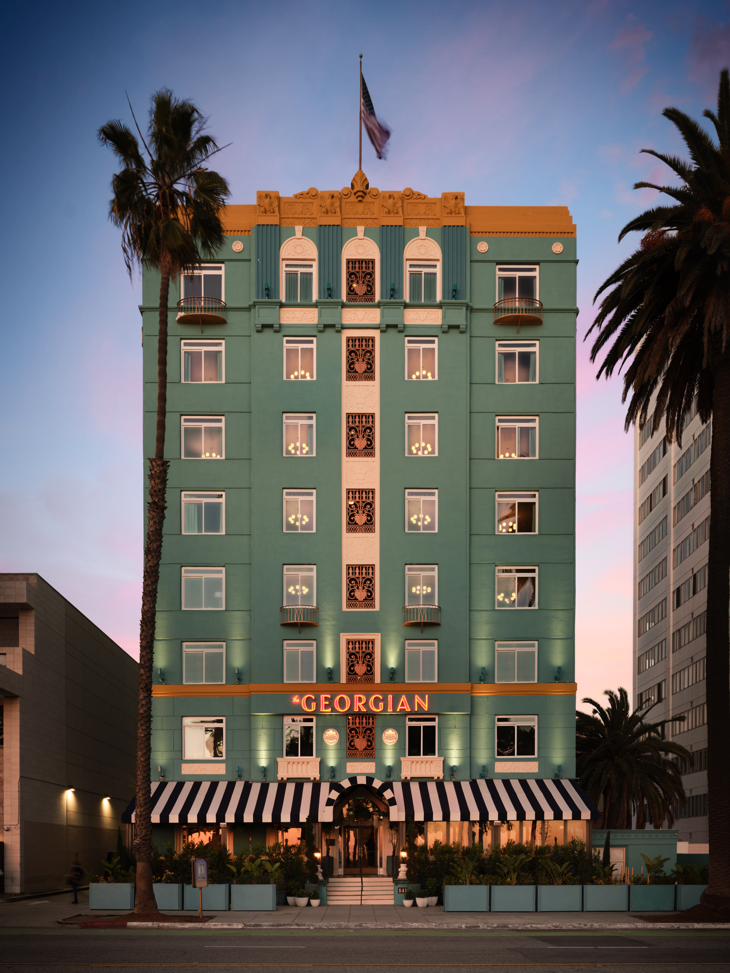 Georgian Hotel in a vertical orientation, showcasing more of its height. The upper floors, adorned with ornate details and ironwork against the teal walls, stand out under the soft sky, while a softly waving flag tops the building.