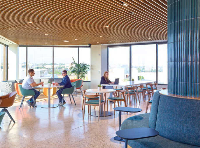 Spacious, sunlit lounge area with floor-to-ceiling windows featuring terrazzo flooring, mid-century modern furniture, and an acoustic fabric-wrapped column. Suspended wooden slats create a warm, textured ceiling, complementing the room's earth-toned color palette and offering a serene work and social environment.