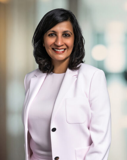 A portrait of a woman with a pleasant smile, medium-length dark hair, and a professional demeanor. She's dressed in a light pink blazer over a matching outfit, standing against a blurred background that suggests a corporate or business setting.