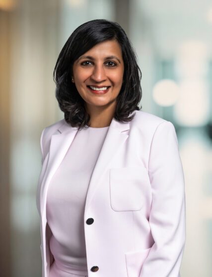 A portrait of a woman with a pleasant smile, medium-length dark hair, and a professional demeanor. She's dressed in a light pink blazer over a matching outfit, standing against a blurred background that suggests a corporate or business setting.