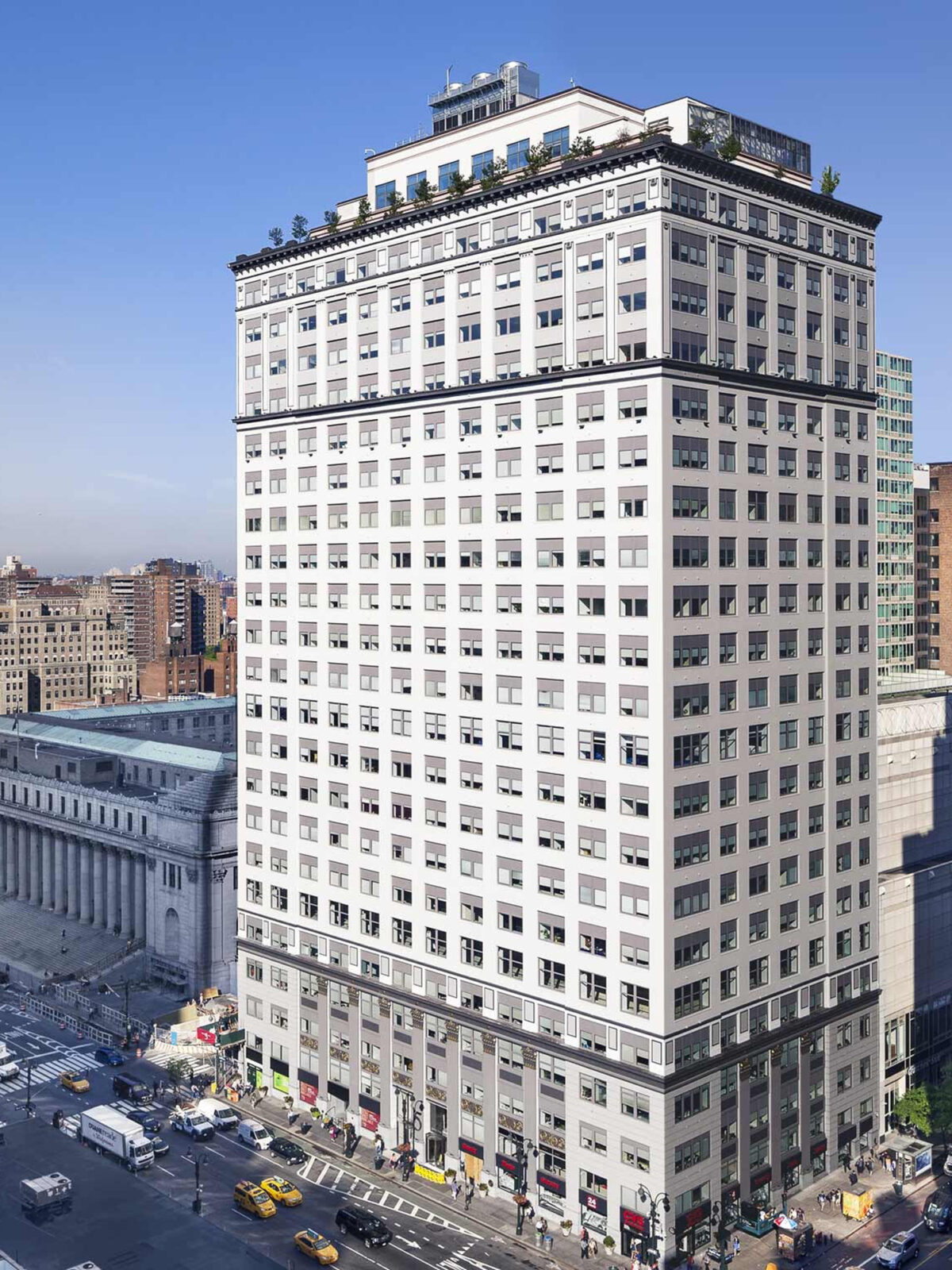 The image features a striking, classic high-rise building, its detailed façade crowned with a rooftop garden, standing prominently at an urban intersection bustling with vehicles and pedestrians, under a clear blue sky.