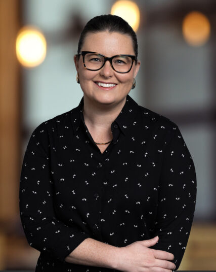 A smiling woman with shoulder-length dark hair poses for a professional portrait. She is wearing large black-framed glasses, a black blouse with a small white pattern, and small hoop earrings, conveying a friendly and approachable demeanor.