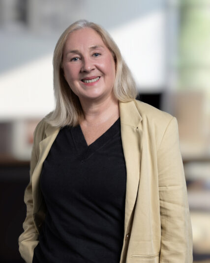 A professional portrait of a woman with long, straight blonde hair, smiling gently at the camera. She is wearing a beige blazer over a black top, and her relaxed yet confident pose in an office setting suggests experience and authority.