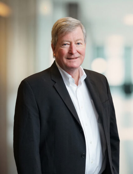 This image shows a portrait of a senior Caucasian man standing in an office environment. He has short, light brown hair, and is smiling gently at the camera. He is wearing a black blazer over a white dress shirt. The background is softly blurred with natural light filtering through large windows, suggesting a professional setting.