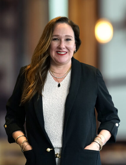The image shows a woman with long dark hair and a beaming smile, wearing a black outfit with a patterned scarf and layered necklaces. The backdrop is softly focused with light tones, suggesting an open and contemporary workspace.