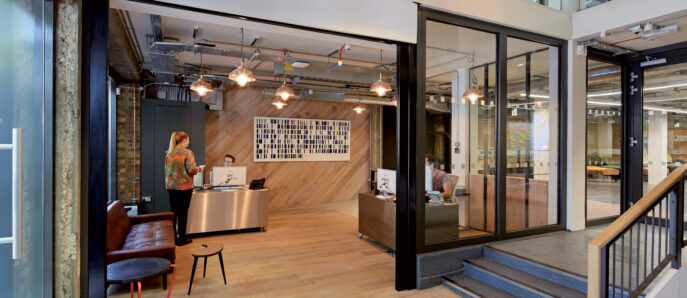 Modern open-plan office space blending industrial and contemporary styles, featuring exposed ductwork, concrete flooring, and pendant lighting. Glass partitions provide transparency while maintaining distinct work areas, complemented by wooden accents and a welcoming reception desk.