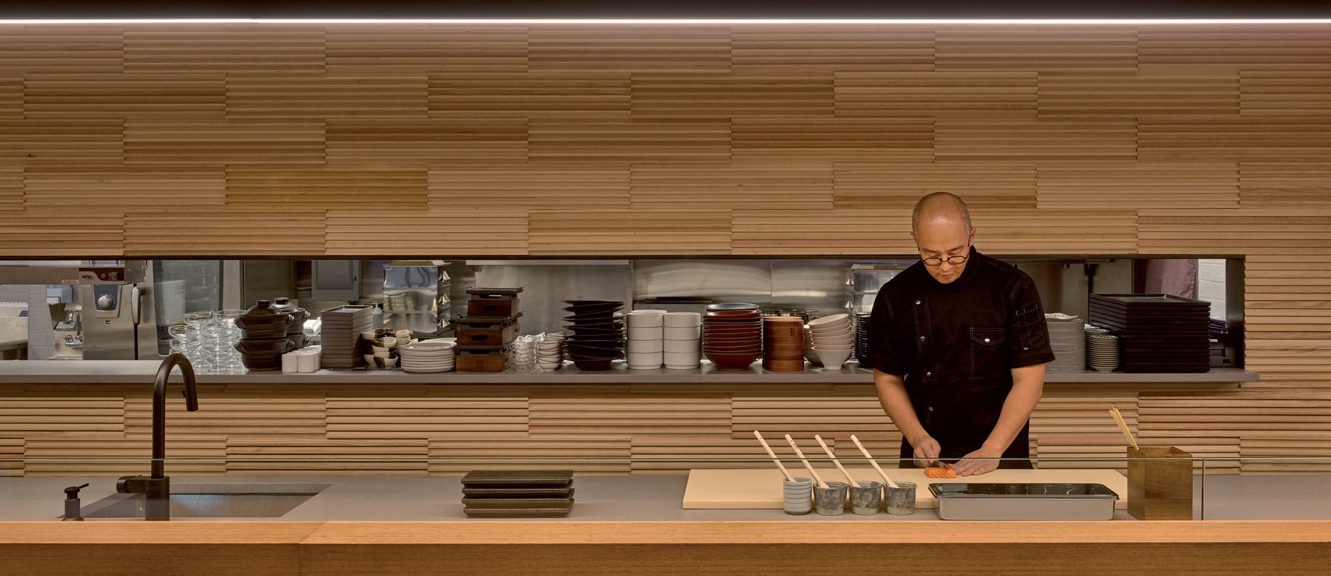 Chef meticulously prepares food at a sleek restaurant kitchen counter, framed by warm wooden slat wall panels that create a rhythmical texture, complemented by minimalist plateware and ambient lighting that casts a cozy glow over the culinary scene.