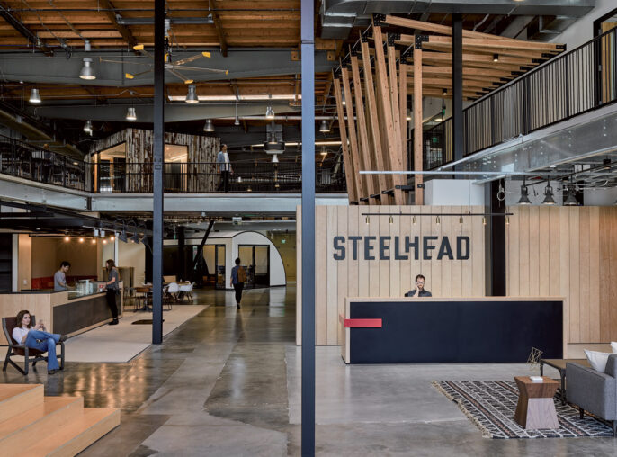 Spacious industrial-style office interior featuring exposed wooden beams, HVAC ductwork, and pendant lighting. The open-plan layout includes a mezzanine level, modern furnishings, and a muted color palette that accentuates the natural wood and metalwork. The Steelhead branded reception area anchors the design.