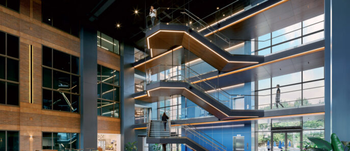 Spacious lobby with high ceiling, illuminated by an angular modern chandelier. Central floating staircase with glass balustrades and wood accents. Comfortable seating areas feature a mix of red armchairs and gray sofas. Large windows allow natural light to complement the warm interior tones.