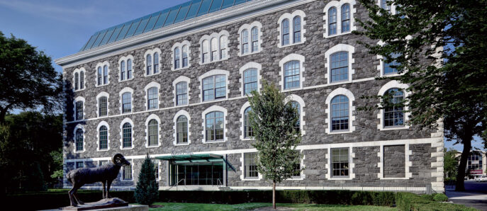 Exterior of a renovated historical building, showcasing a well-preserved stone facade, symmetrically aligned arched windows, and a modern glass rooftop addition that contrasts with the traditional architecture below. A sculptural art piece adorns the manicured front lawn.