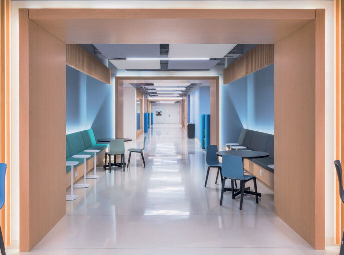 Modern corridor with clean lines, featuring wood-accented alcoves, teal and blue furnishing, and efficient LED lighting guiding the way. The design merges functionality with comforting aesthetics to ease navigation and enhance patient comfort.