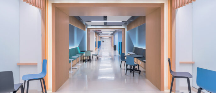 Modern corridor with clean lines, featuring wood-accented alcoves, teal and blue furnishing, and efficient LED lighting guiding the way. The design merges functionality with comforting aesthetics to ease navigation and enhance patient comfort.