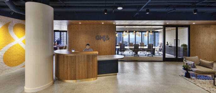 Modern office reception area blending organic and industrial motifs, with a curved wood-paneled desk, sleek pendant lighting, exposed ceiling features, and a monogram logo accent wall. Brickwork adds texture while the adjacent conference room showcases sleek, transparent partitions.