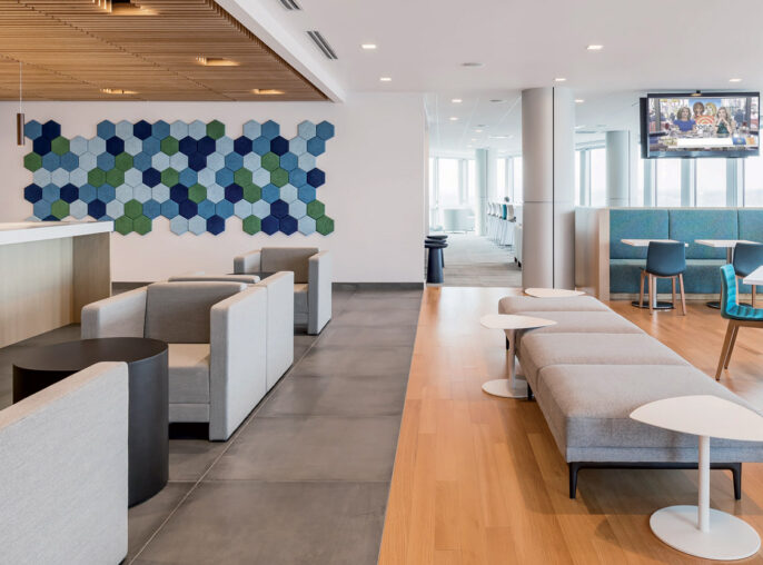 Modern office breakroom featuring natural light, sleek white surfaces, wooden slats ceiling detail, and pops of color from blue upholstered furniture and hexagonal wall art.