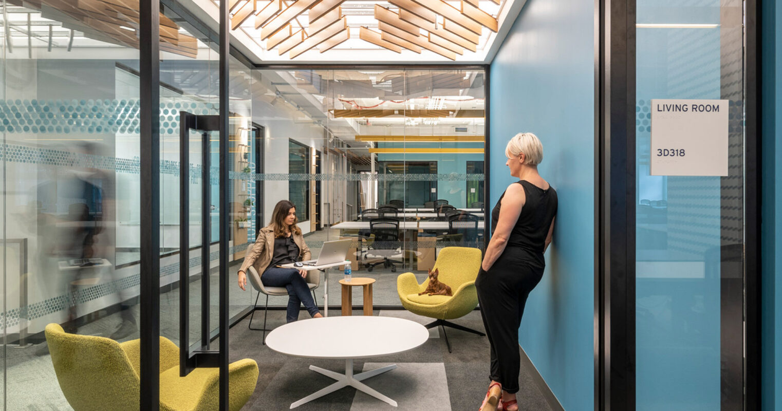 Modern office breakout area with glass walls, featuring a wood slat ceiling that cascades down as wall accent. Two individuals occupy the vibrant space, one seated at a round table on chartreuse chairs, the other walking through, reflecting dynamic workplace design fostering both interaction and movement.