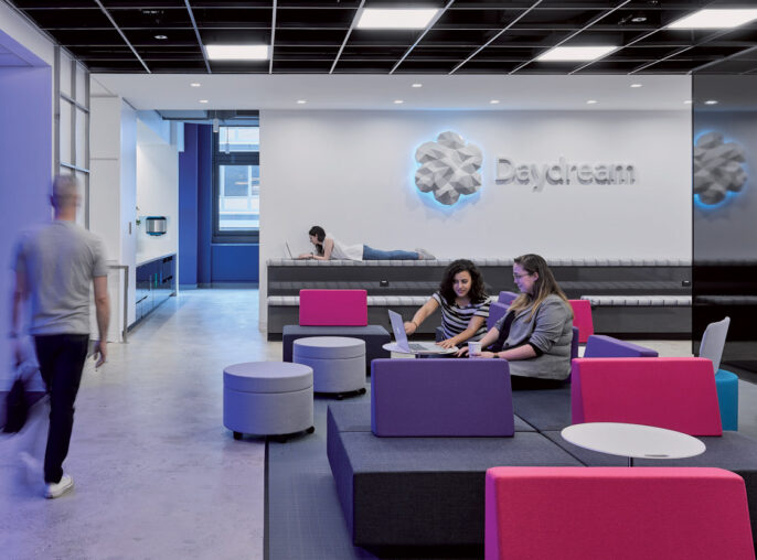 Modern office lounge featuring blue and magenta furniture with clean lines, accented by dynamic geometric carpet patterns. A boldly branded wall provides a backdrop to collaborative seating areas, emphasizing open-concept design and facilitating informal teamwork.