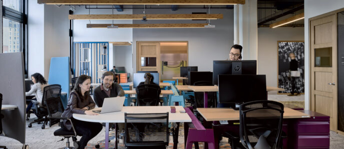 Modern open-plan office space featuring eclectic furniture with a combination of communal desks, private booths, and a lounge area, accented by vibrant carpets and exposed ceiling beams. Employees are engaged in collaborative and individual work under ample natural and ambient lighting.