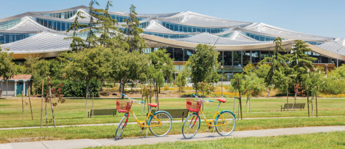 Lush green lawn fronts a modern architectural marvel with undulating glass facade and white supports, flanked by vibrant yellow bicycles in sunny park-like setting.