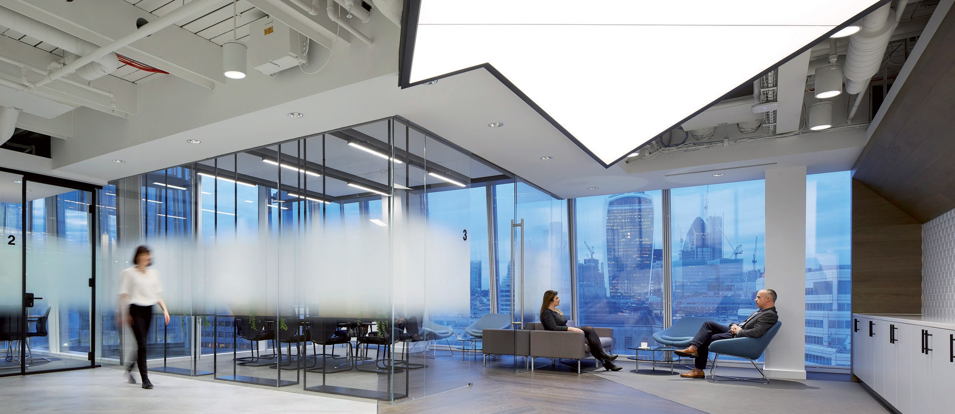 Modern office space featuring floor-to-ceiling glass partitions, sleek furnishings, and a neutral color palette. Ambient lighting complements the natural light flooding in from exterior windows, offering views of the urban skyline. Dynamic wood flooring contrasts with the structured ceiling design.