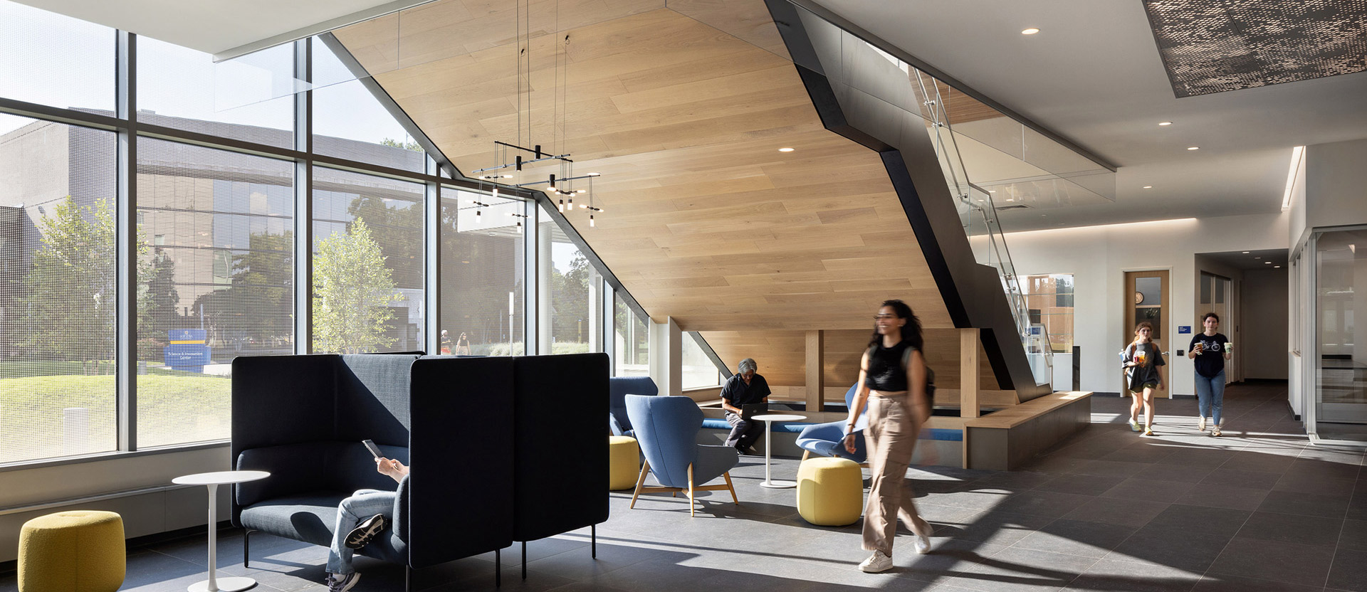 Warm natural light floods a modern lobby with a geometric wood-paneled staircase as the centerpiece. Sleek, dark furniture contrasts with the light floor, accented by vibrant yellow ottomans. Expansive glass walls offer a seamless indoor-outdoor connection.