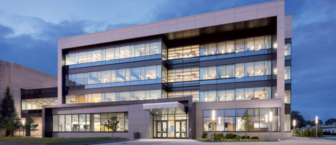 Modern architectural design featuring a multi-level office building with illuminated windows against twilight sky, showcasing reflective glass facades and geometric symmetry with an emphasis on horizontal lines.