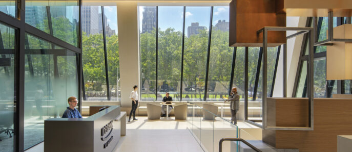 Modern office lobby with floor-to-ceiling windows offering an expansive view of greenery, bathed in natural light. A sleek reception desk serves as focal point, flanked by geometric wooden design elements and casual seating areas for visitors.