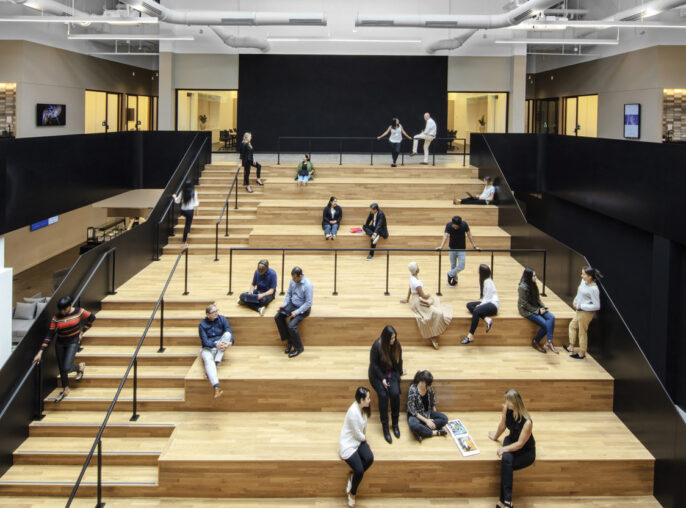 Spacious, modern interior features tiered wooden seating with people casually scattered, engaging in discussions and work. Above, a white industrial ceiling with exposed ductwork contrasts starkly with the warm-toned flooring and matte black partitions, highlighting a contemporary, collaborative workspace design.