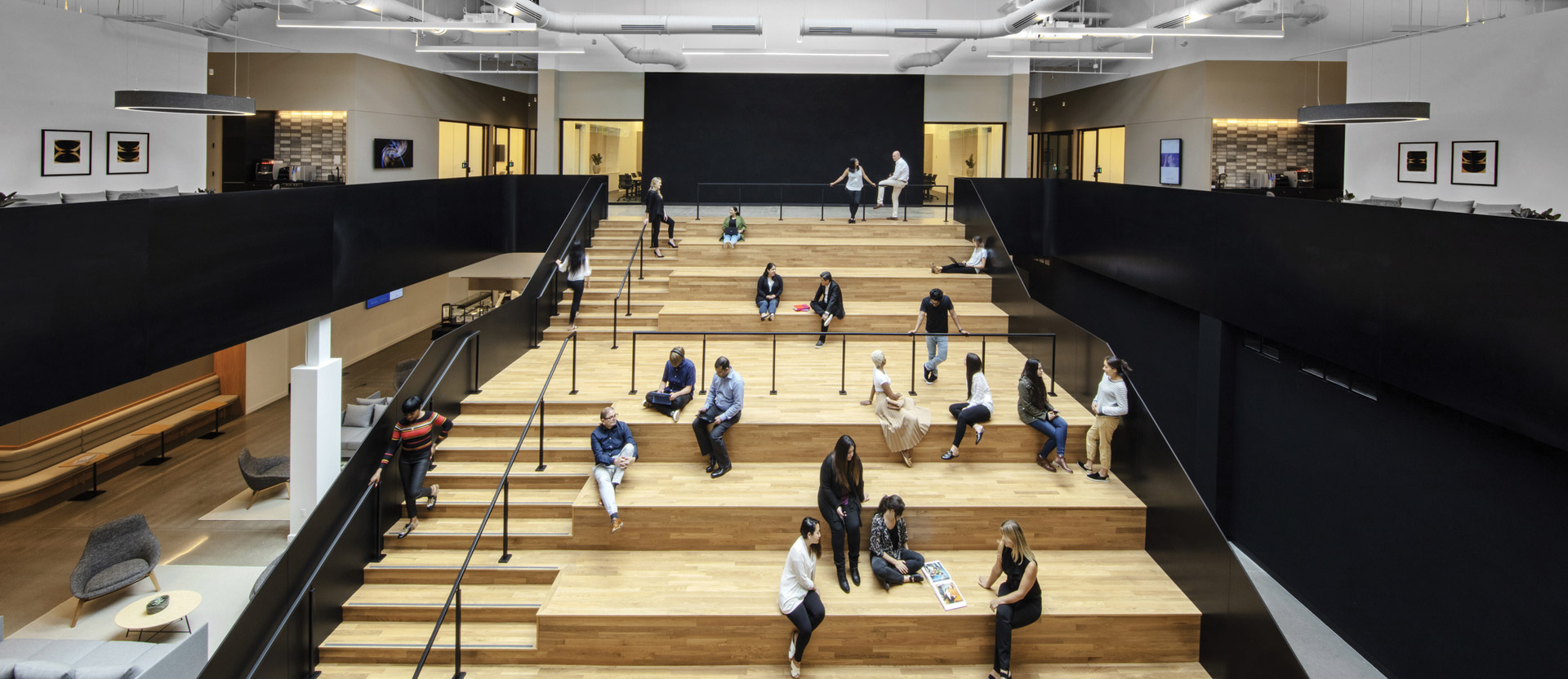 Spacious, modern interior features tiered wooden seating with people casually scattered, engaging in discussions and work. Above, a white industrial ceiling with exposed ductwork contrasts starkly with the warm-toned flooring and matte black partitions, highlighting a contemporary, collaborative workspace design.