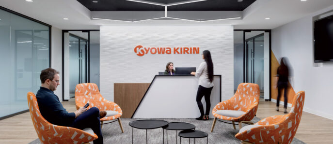 Modern corporate reception area featuring bold Kyowa Kirin brand wall, pops of orange in the upholstered armchairs, sleek black center tables, and dynamic lighting overhead, creating a welcoming ambiance for professionals and guests.