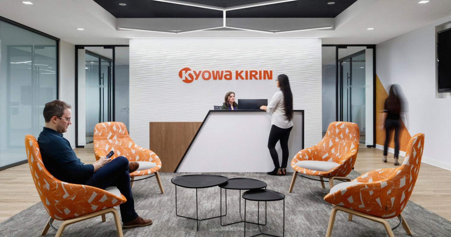 Modern corporate reception area featuring bold Kyowa Kirin brand wall, pops of orange in the upholstered armchairs, sleek black center tables, and dynamic lighting overhead, creating a welcoming ambiance for professionals and guests.