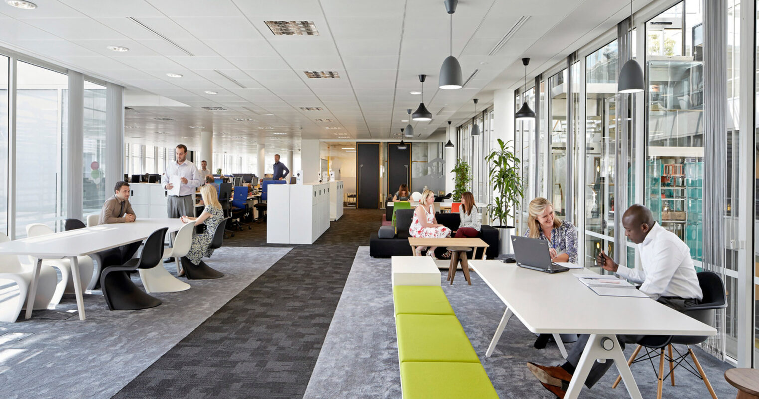 Modern open-plan office space with natural light, featuring an eclectic mix of communal work tables, private cubicles, and casual seating areas. Bright color accents punctuate the neutral color palette, enhancing the environment's energy and fostering a collaborative atmosphere.