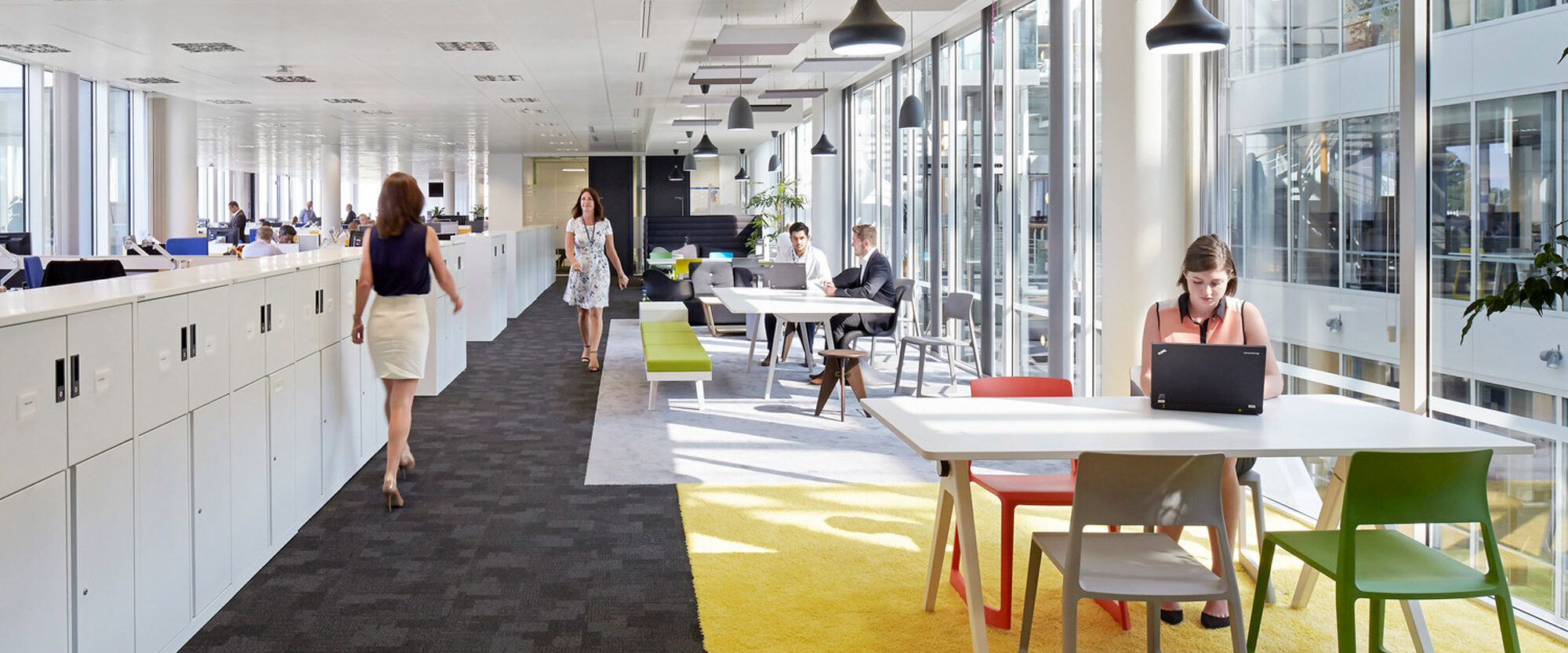 Sunlight streams into a modern office space featuring long white desks, colorful ergonomic chairs, and pendant lighting. Employees work at spaced-out stations promoting productivity and social distancing. The vibrant yellow carpet adds a cheerful accent to the clean, minimalist design.
