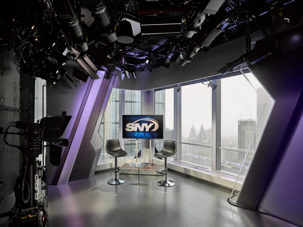 Sleek broadcast studio featuring modern furniture, high-tech lighting, and video equipment with a panoramic window overlooking a cityscape, embodying a contemporary aesthetic.