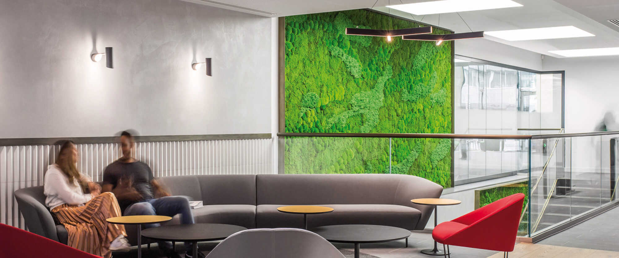 Modern office lounge with a vibrant green living wall feature, sleek gray modular seating, red accent chairs, and unique petal-shaped coffee tables, under linear pendant lighting, on light wood flooring.