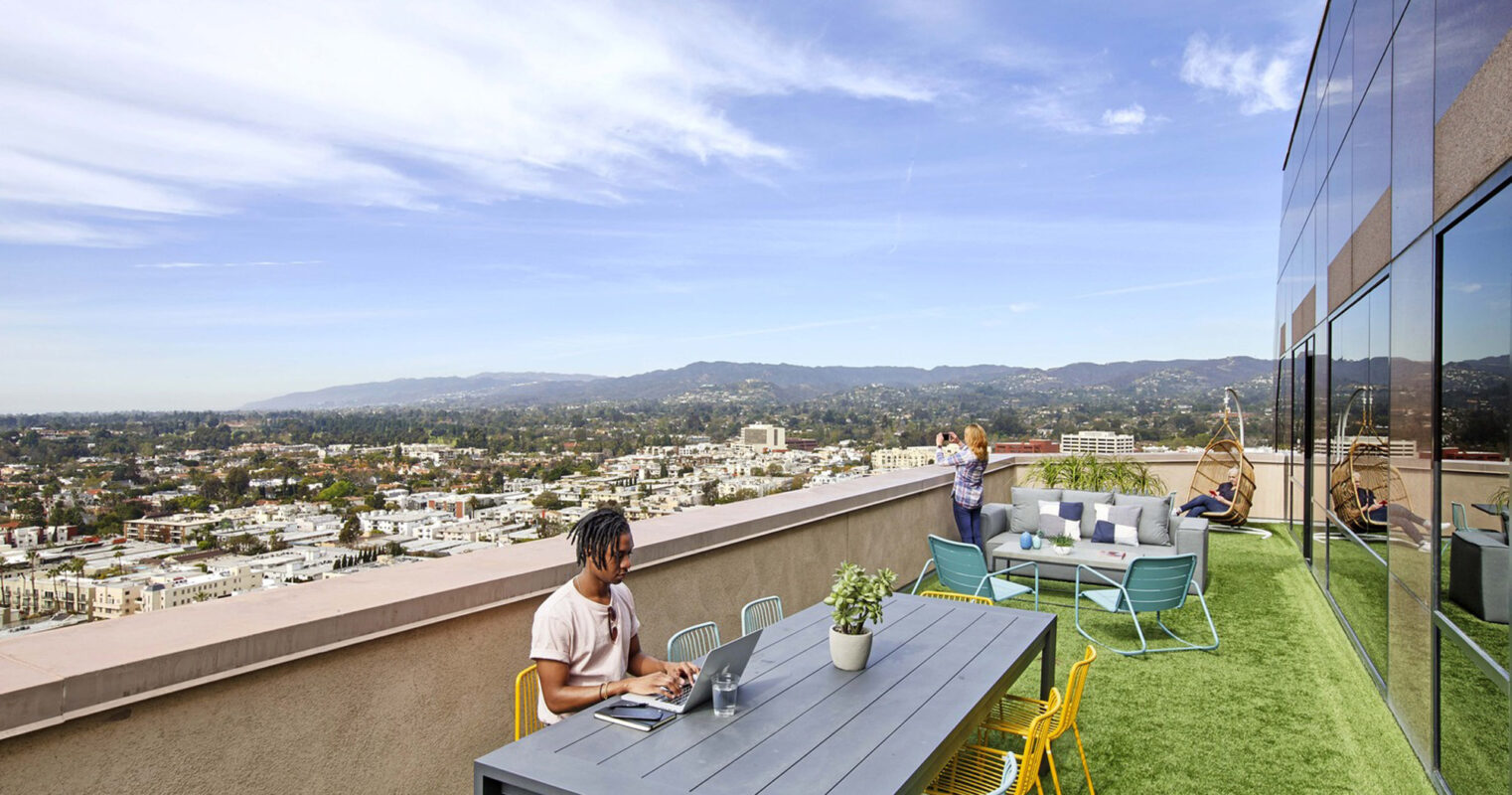 Rooftop terrace with contemporary outdoor furniture, artificial turf, and glass balustrades, offering a panoramic view of an urban skyline. People are seen utilizing the space for work and relaxation amidst potted plants, accentuating the blend of functionality and aesthetics.