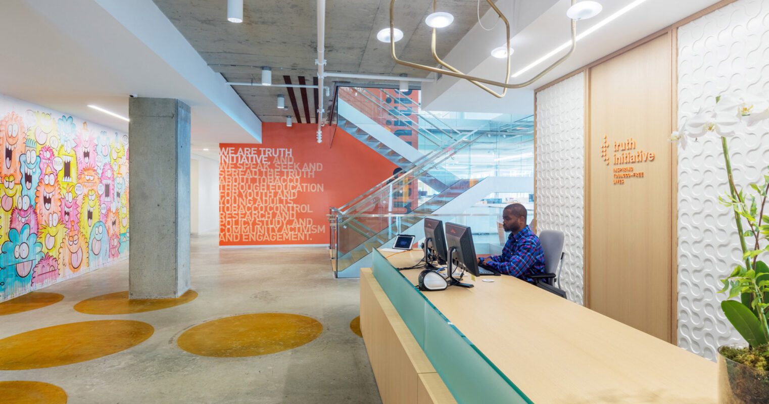 Modern office space blending functionality and art, featuring a vibrant graffiti wall, exposed concrete columns, and a minimalist reception desk with a sleek computer setup. Circular organic floor patterns complement the unique pendant lighting above.