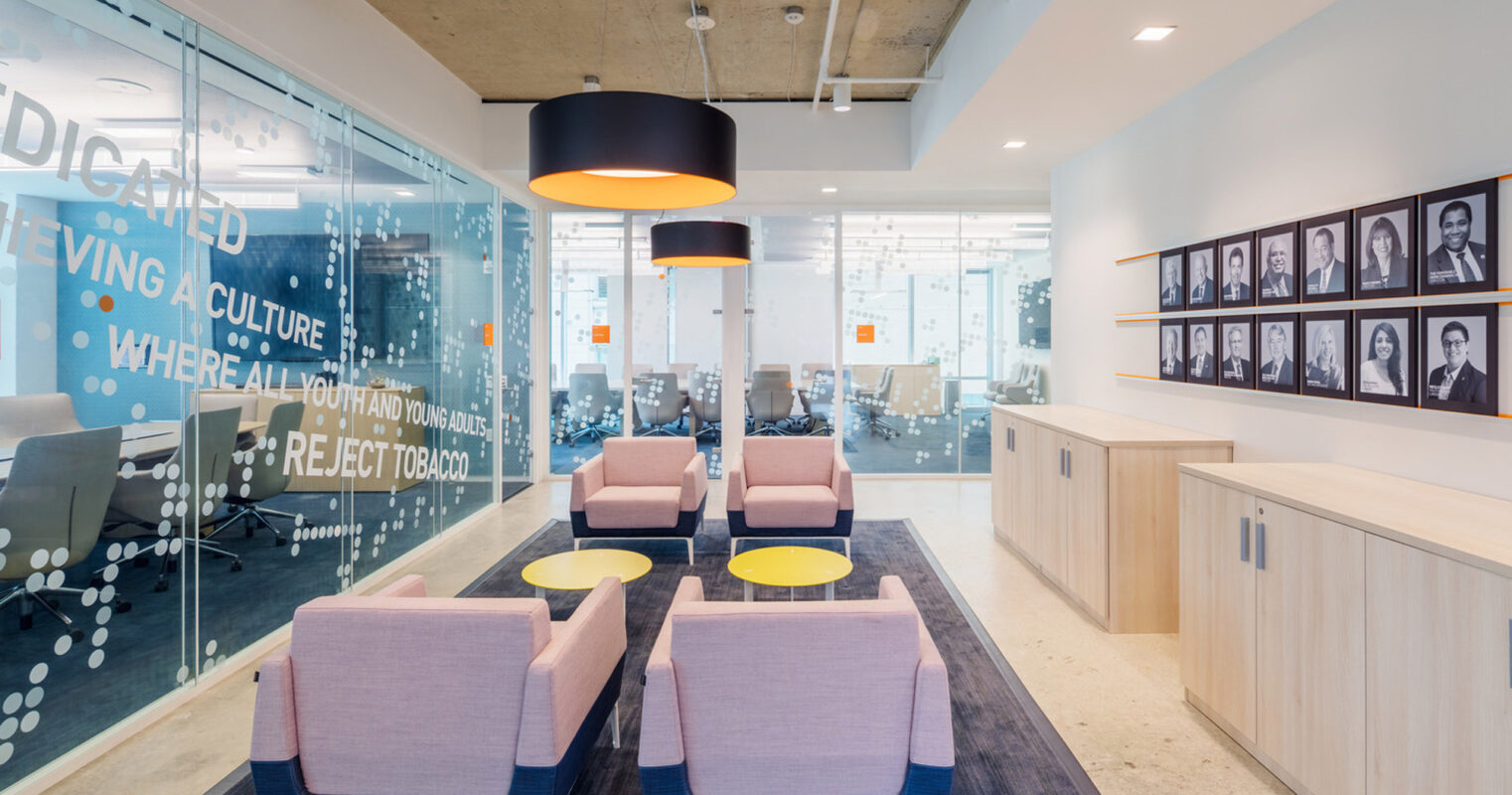 Modern office lounge with pastel pink armchairs centered around a low-profile circular coffee table, with bold circular overhead lighting complementing the space's open-concept design. The expansive glass partition features typographical branding, fostering a collaborative environment amidst monochrome portraits of thought leaders.