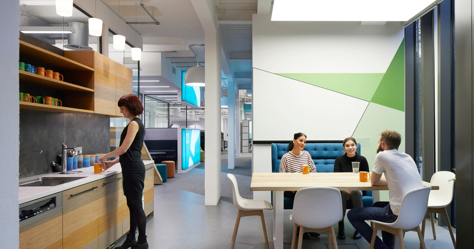 Modern office kitchenette blends form and function with sleek white countertops, vibrant geometric wall accents, and designer pendant lighting, facilitating casual employee interactions in a bright, contemporary space.