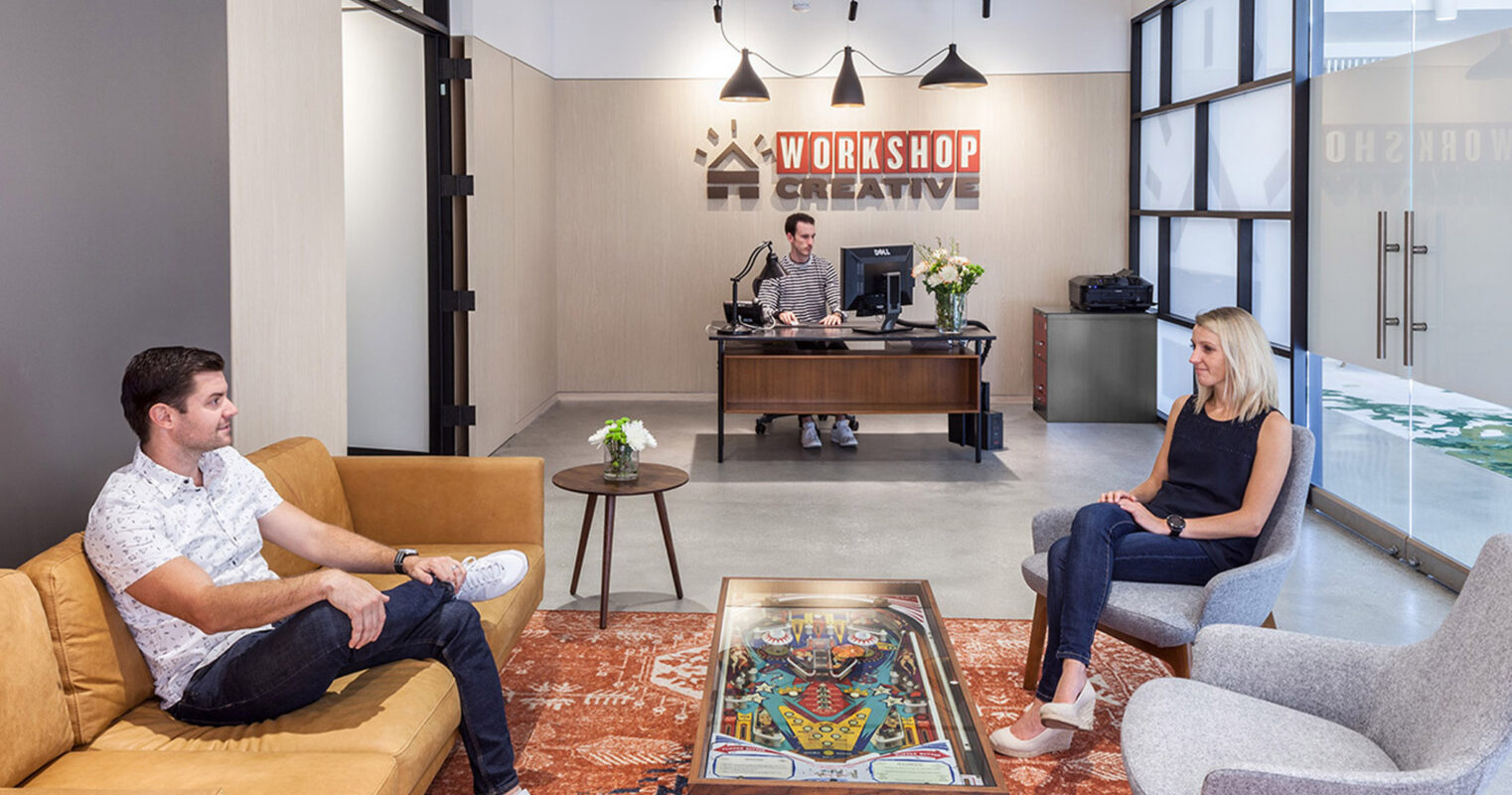 Modern office lobby with a warm color scheme featuring tan leather couches, gray accent chairs, and a pinball coffee table. The reception desk has a backlit 'WORKSHOP' sign. Natural light streams in, complementing the sleek, industrial-chic design elements.