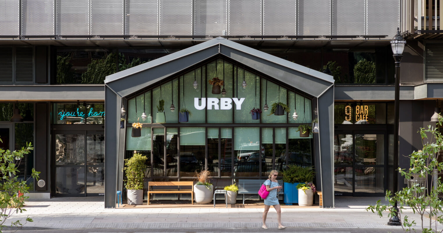 Modern urban cafe exterior with symmetrical design, featuring warm interior lighting, hanging plants, and a welcoming bench under a canopy, flanked by potted greenery.